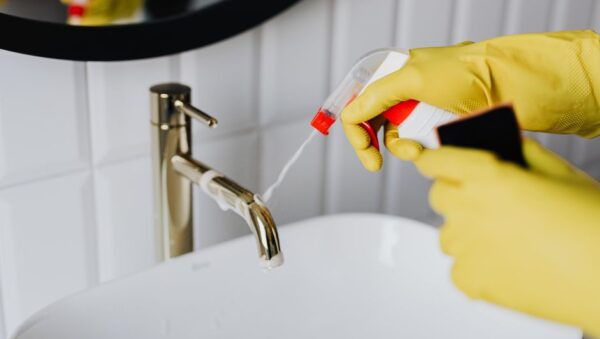 Persona limpiando la cal de un grifo de baño con guantes y spray desincrustante